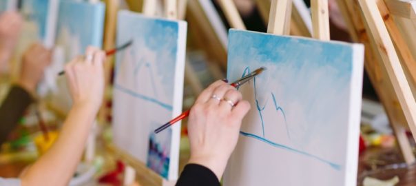 People sitting at painting easels as they paint blue mountains and a sky.
